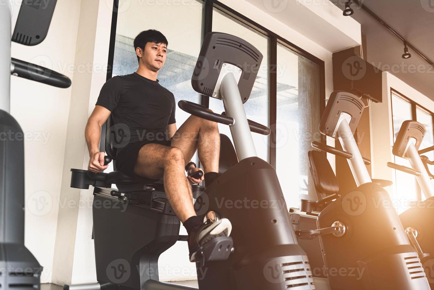 Hombre joven asiático del deporte que monta la bicicleta estacionaria en el gimnasio foto