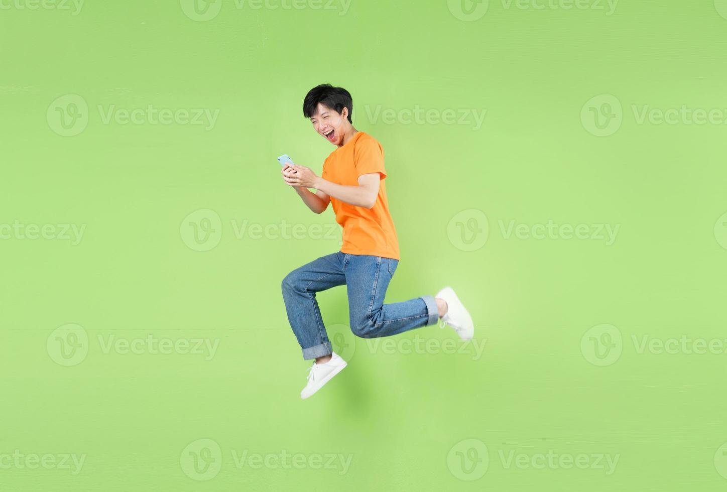 Hombre asiático saltando y sosteniendo el teléfono inteligente, aislado en verde foto