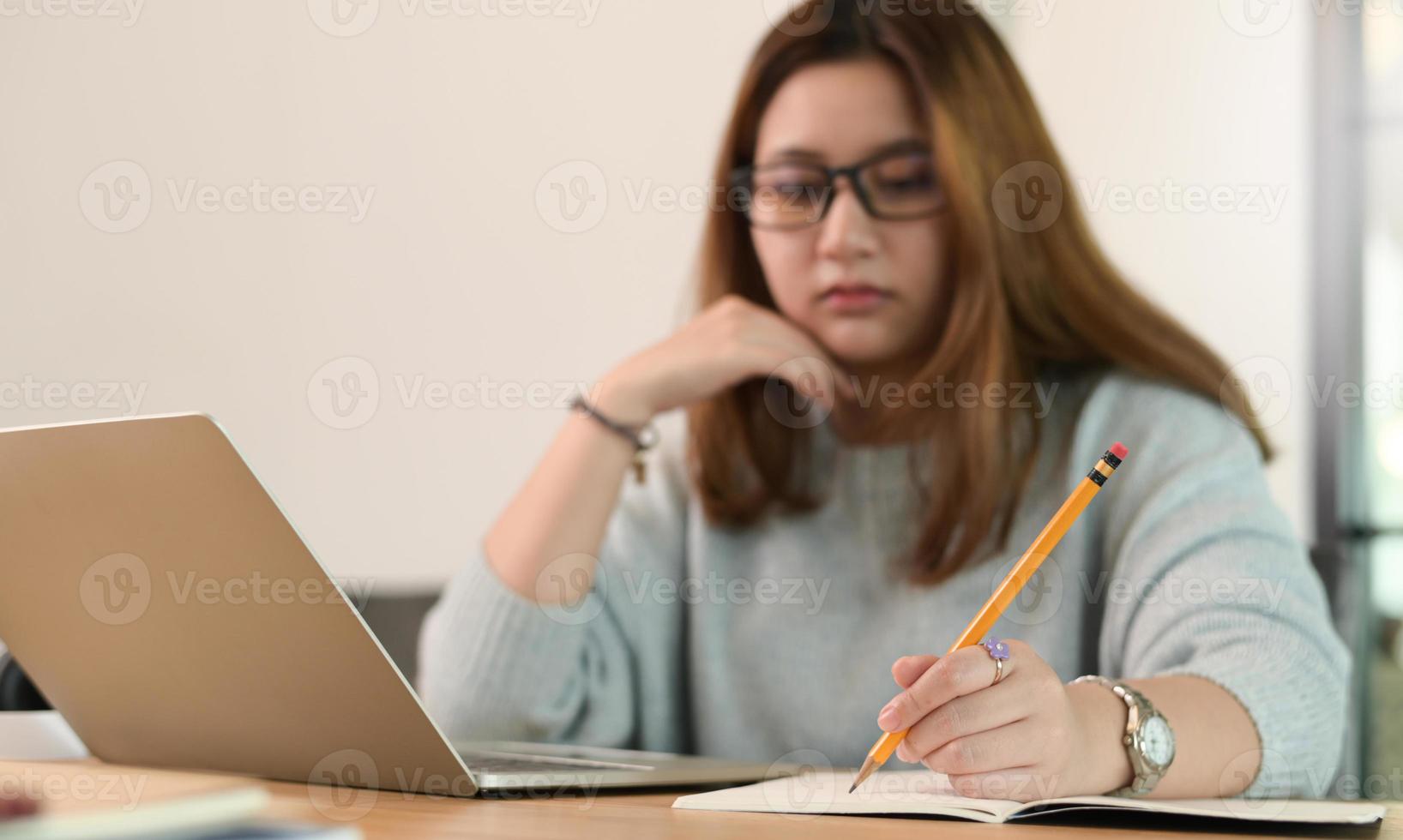 A teenage girl wearing glasses uses a pencil to write on a notebook. photo