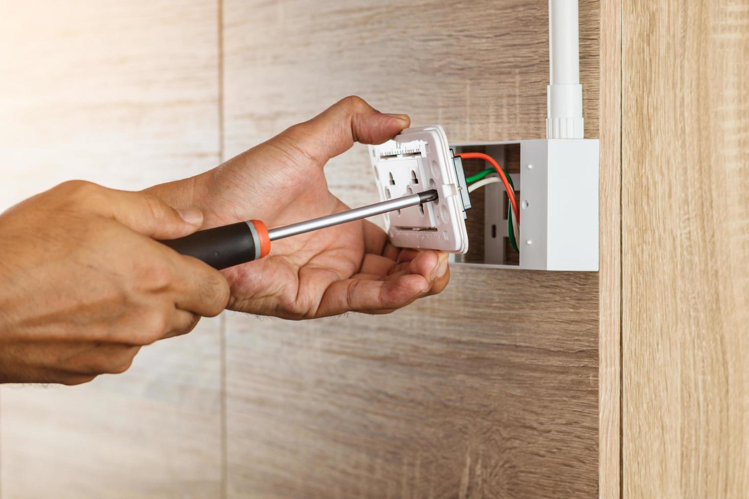 El electricista está usando un destornillador para instalar una toma de corriente en una caja de plástico en una pared de madera. foto