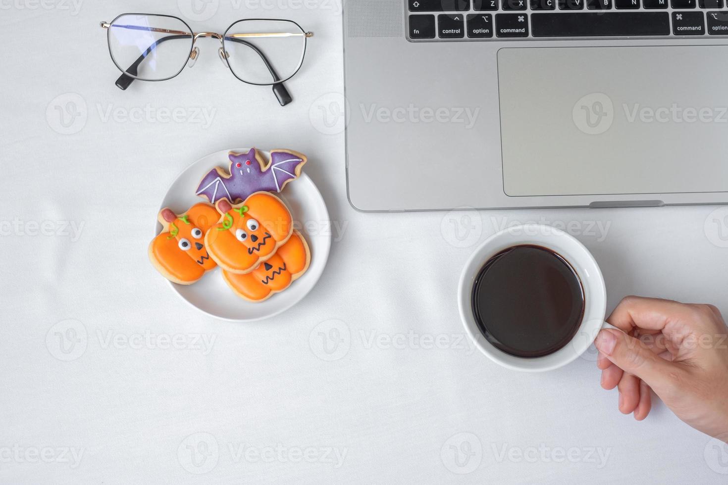 Hand holding coffee cup and eating Halloween Cookies during using computer laptop. Happy Halloween, online shopping, Hello October, fall autumn, Festive, party and holiday concept photo