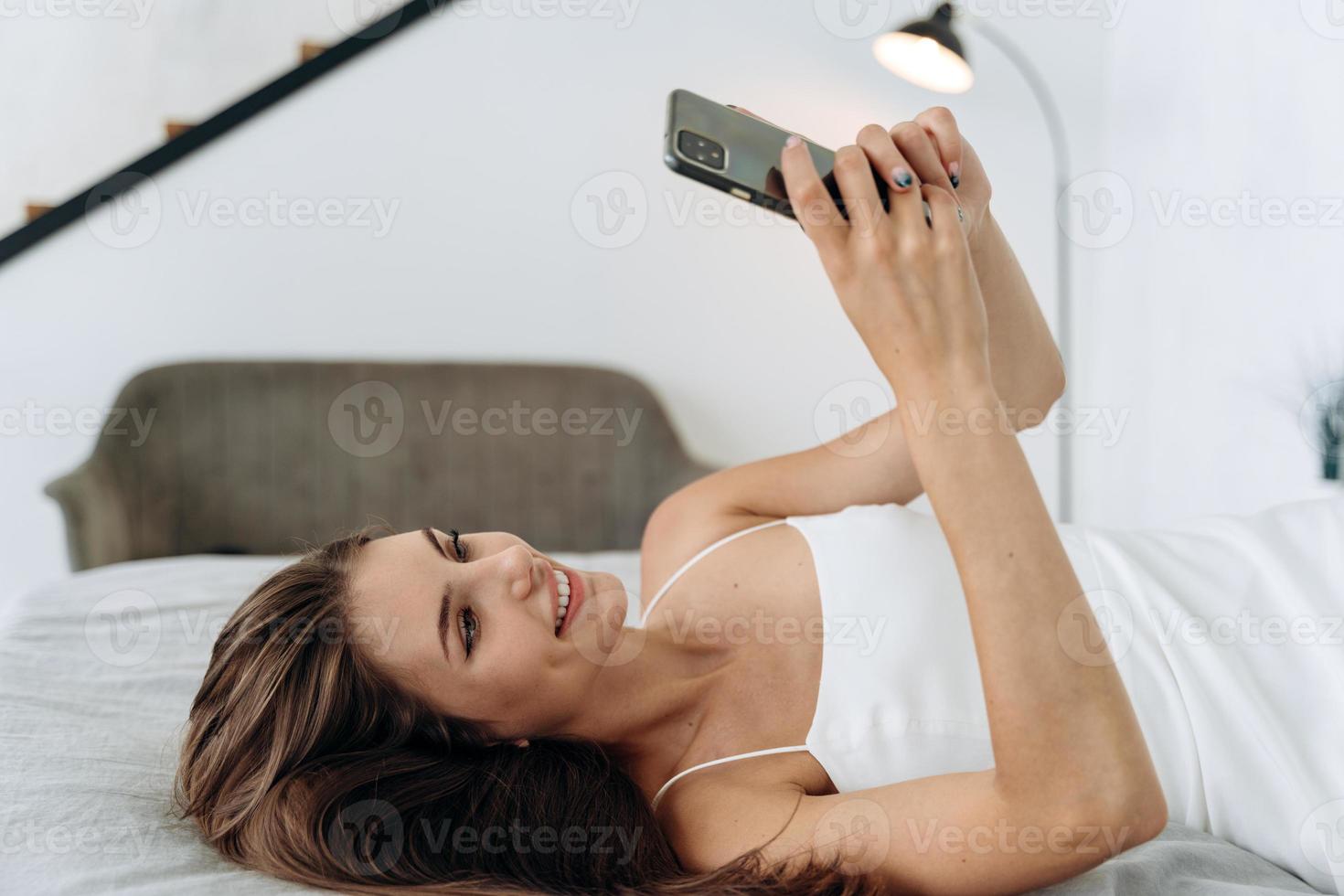 feliz mujer joven morena con teléfono móvil en casa. hermosa chica mirando el teléfono inteligente y navegando por la red. Mujer sonriente mirando el teléfono móvil mientras está acostada en su cama foto