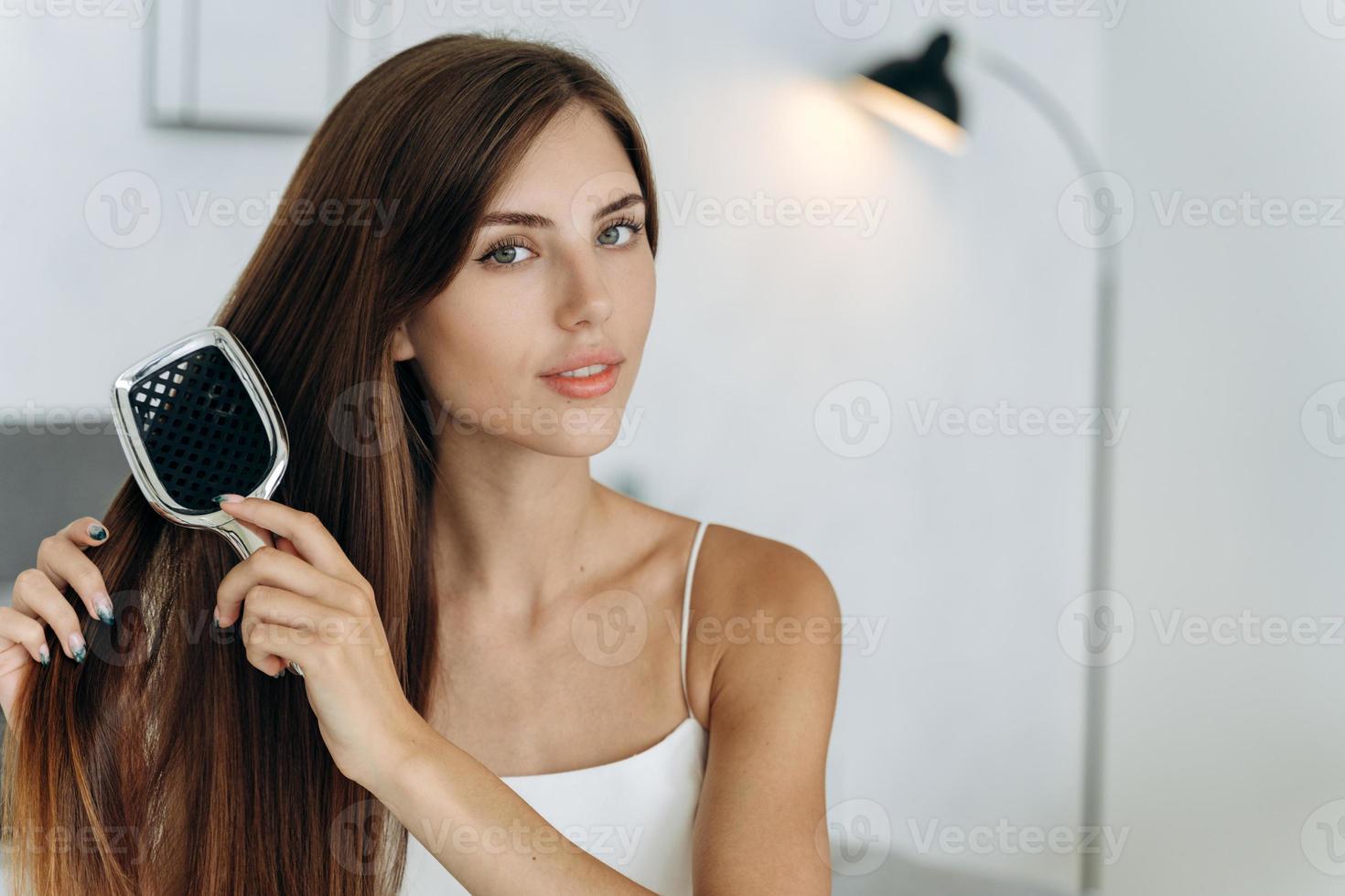 Young confident woman wearing domestic clothes sitting at the bed and brush her long healthy brunette hair. Millennial female doing morning beauty routine photo