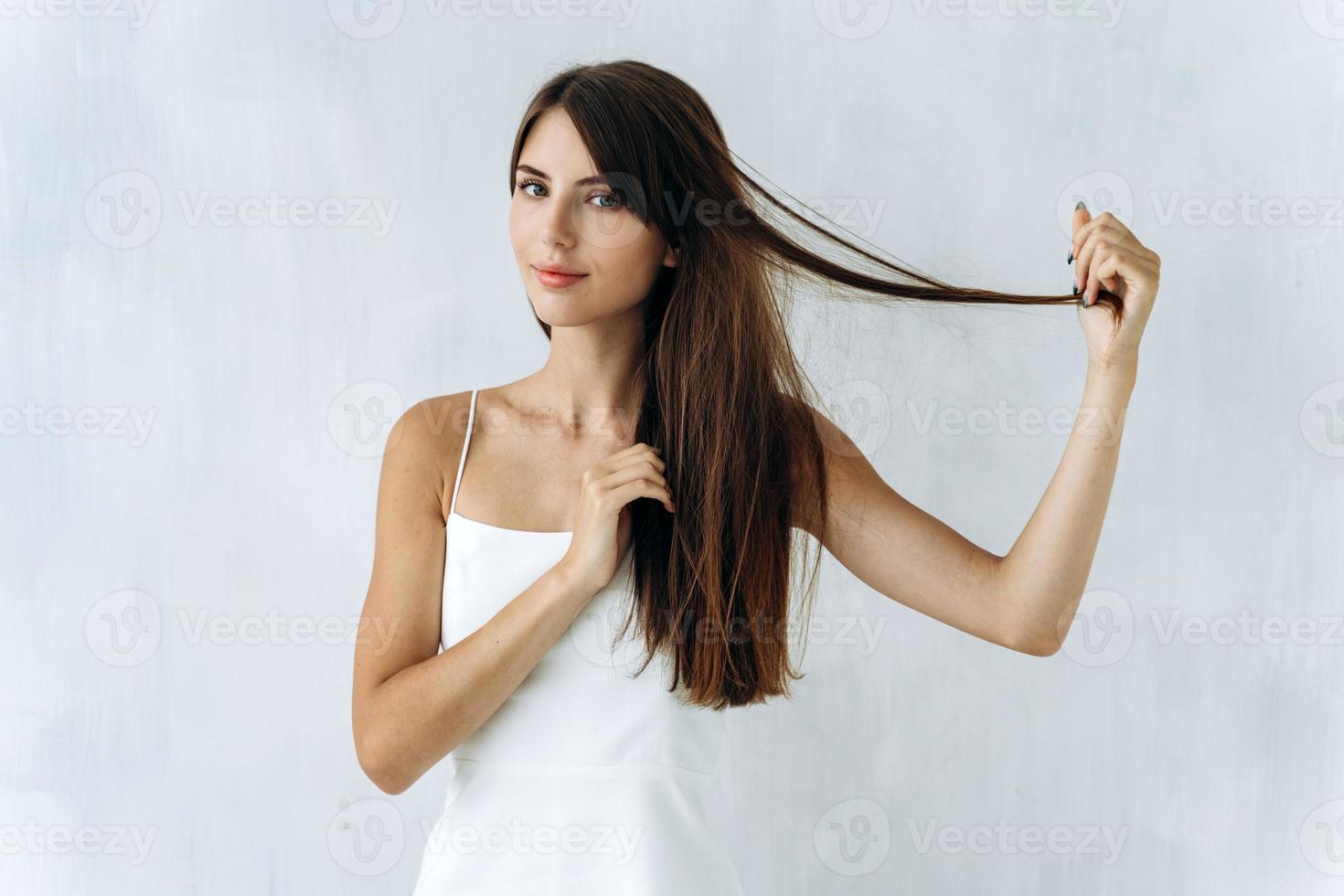 Cintura para arriba retrato de la mujer caucásica de pelo largo poniendo sus manos en su cabello y jugando con él mientras posa con una pared blanca en el fondo foto