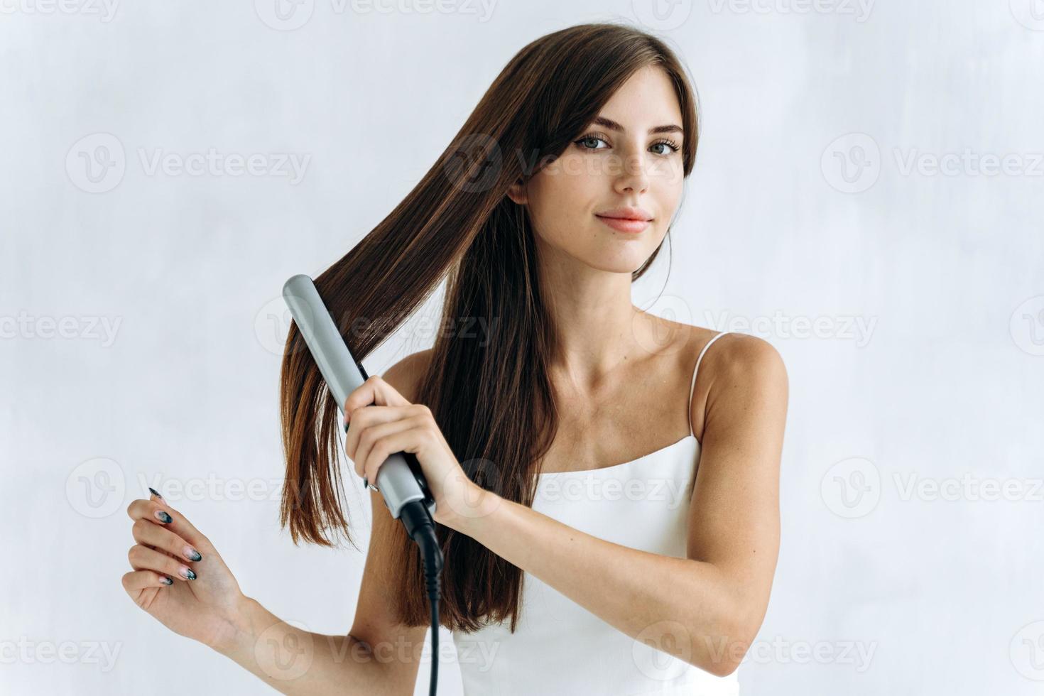 cintura para arriba retrato de la mujer joven contenta con una plancha de pelo en el baño mientras mira a la cámara con una sonrisa de placer. concepto de apariencia de mujer foto