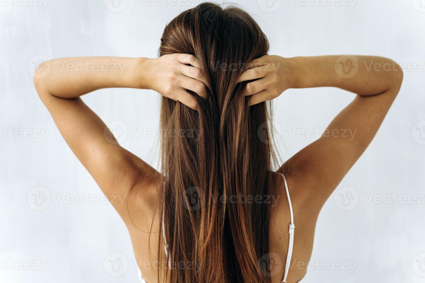 Back view photo of young beautiful woman touching to straight brown hair with her hands isolated on white color background. Woman hair concept