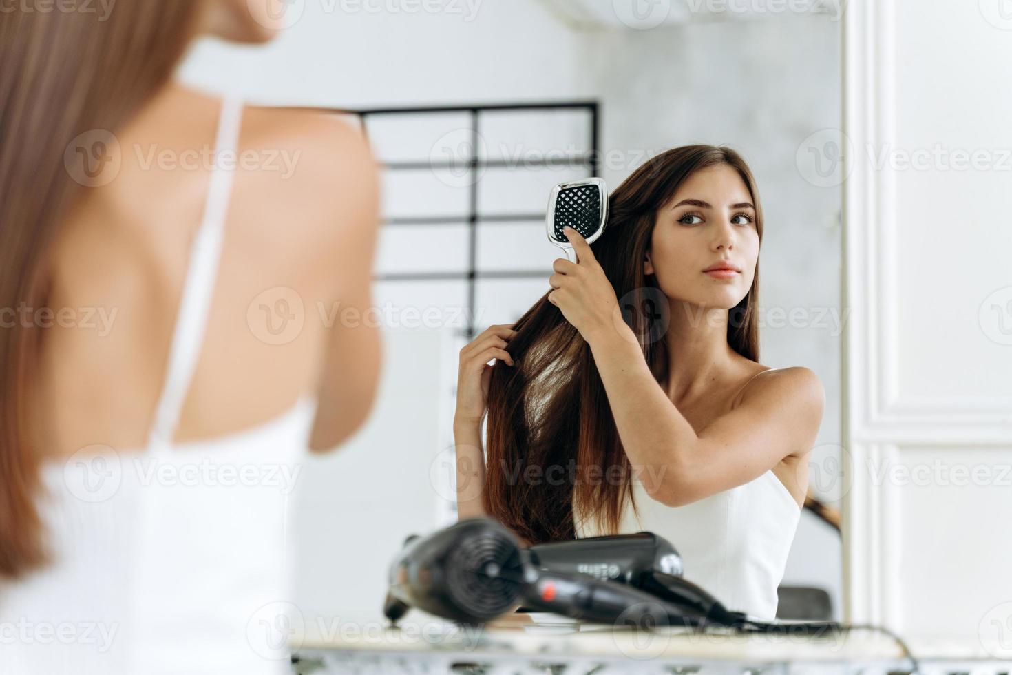 peinado y cuidado diario del cabello en casa. Alegre dama caucásica milenaria peinando largo y hermoso cabello lacio en el interior del baño en la mañana foto