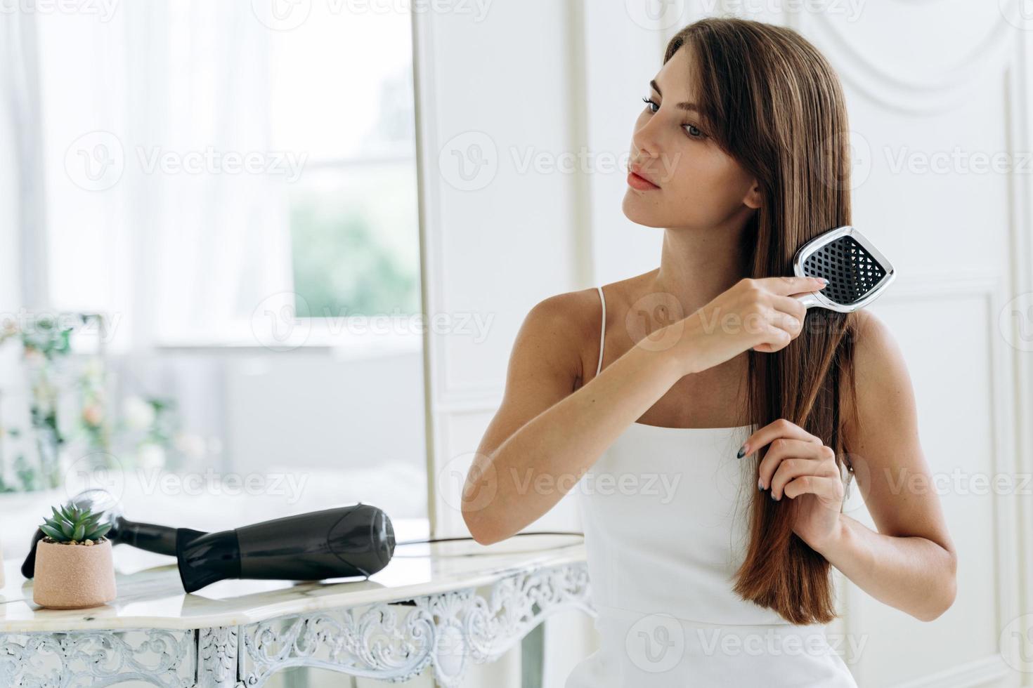 Retrato de cintura para arriba de una dama radiante que se peina el cabello largo y oscuro mientras mira hacia otro lado con una sonrisa tranquila. concepto de cuidado del cabello. foto de stock