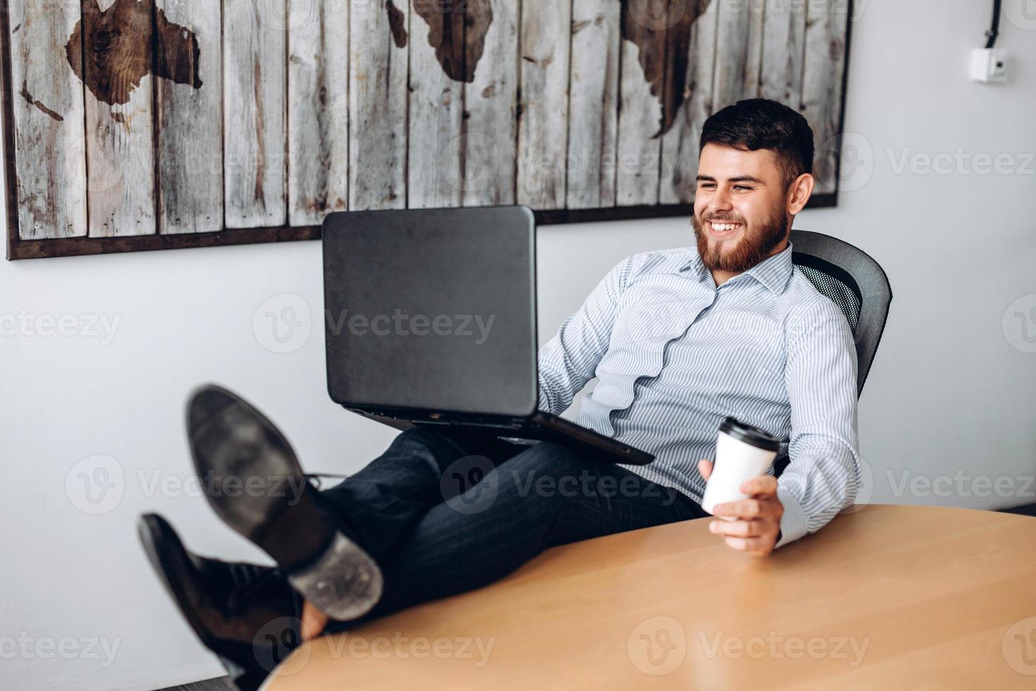 buen chico con barba sentado en una mesa, poniendo los pies sobre la mesa, tomando café y trabajando en una computadora foto