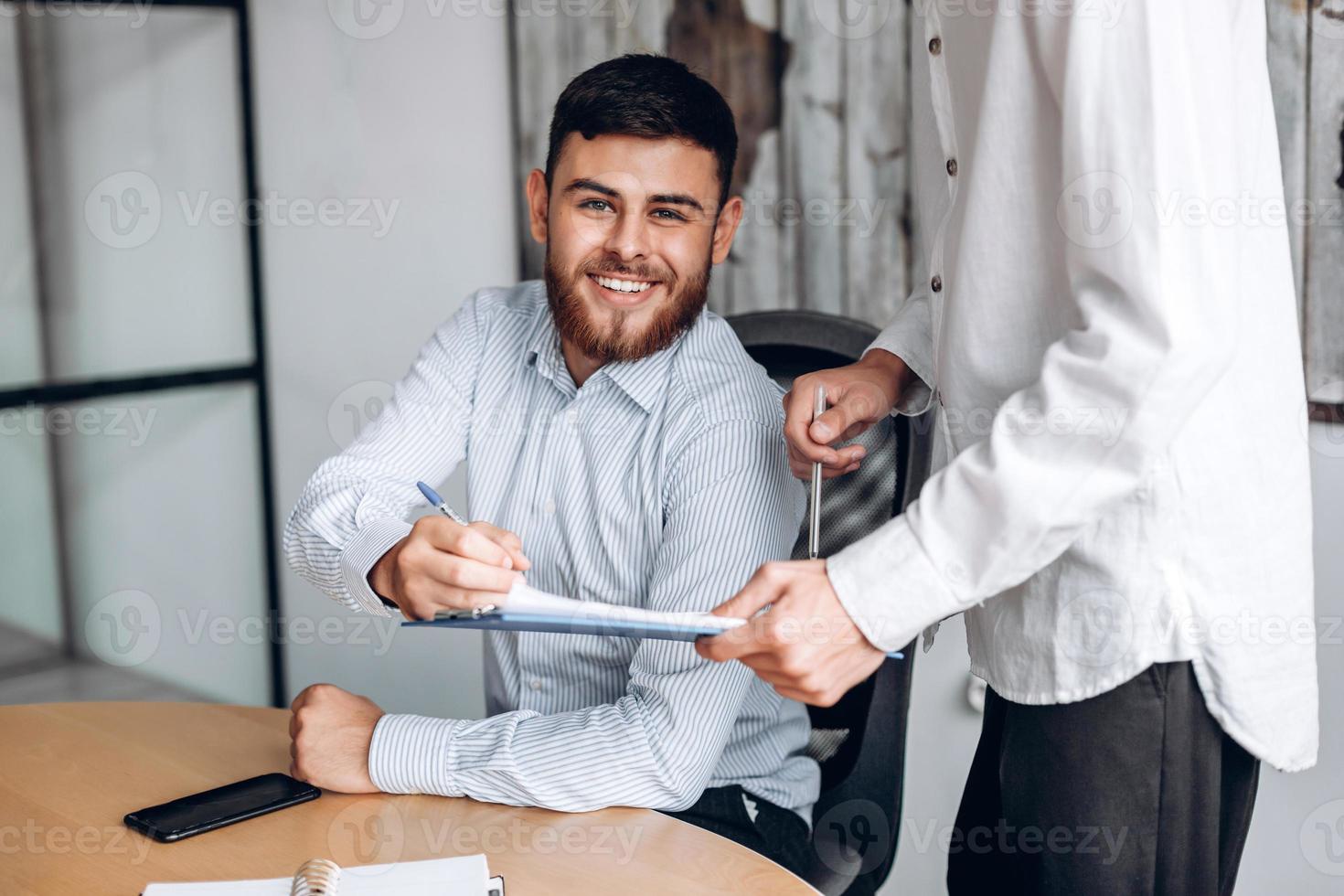 hombre sonriente con barba, trabaja en la oficina, mira documentos importantes y los firma foto