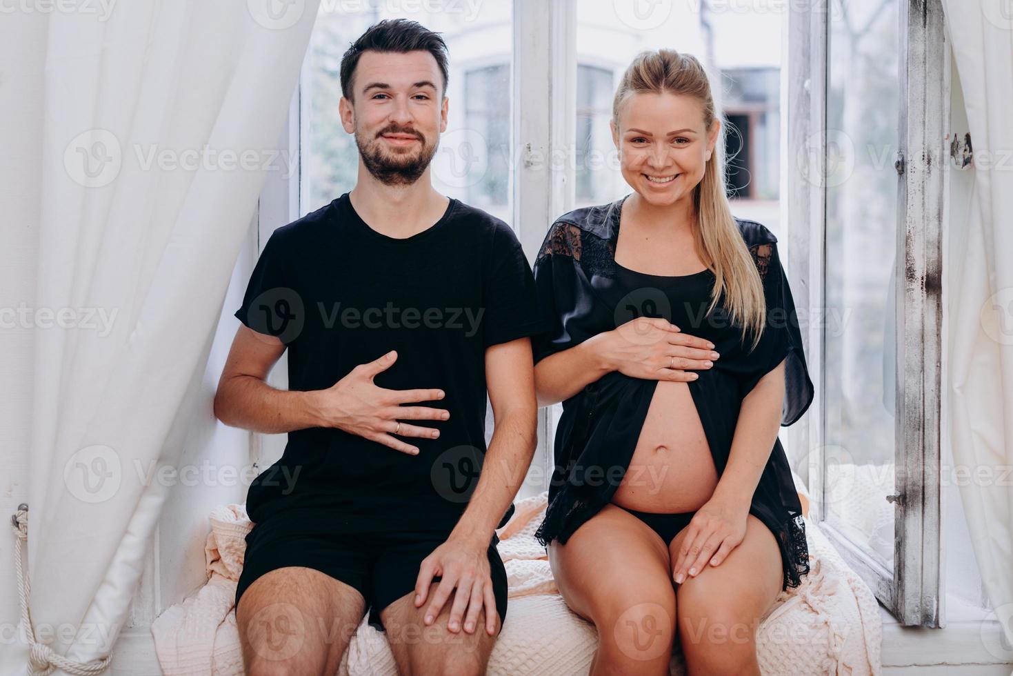 Happy funny couple posing on window and showing belly of pregnant woman. photo