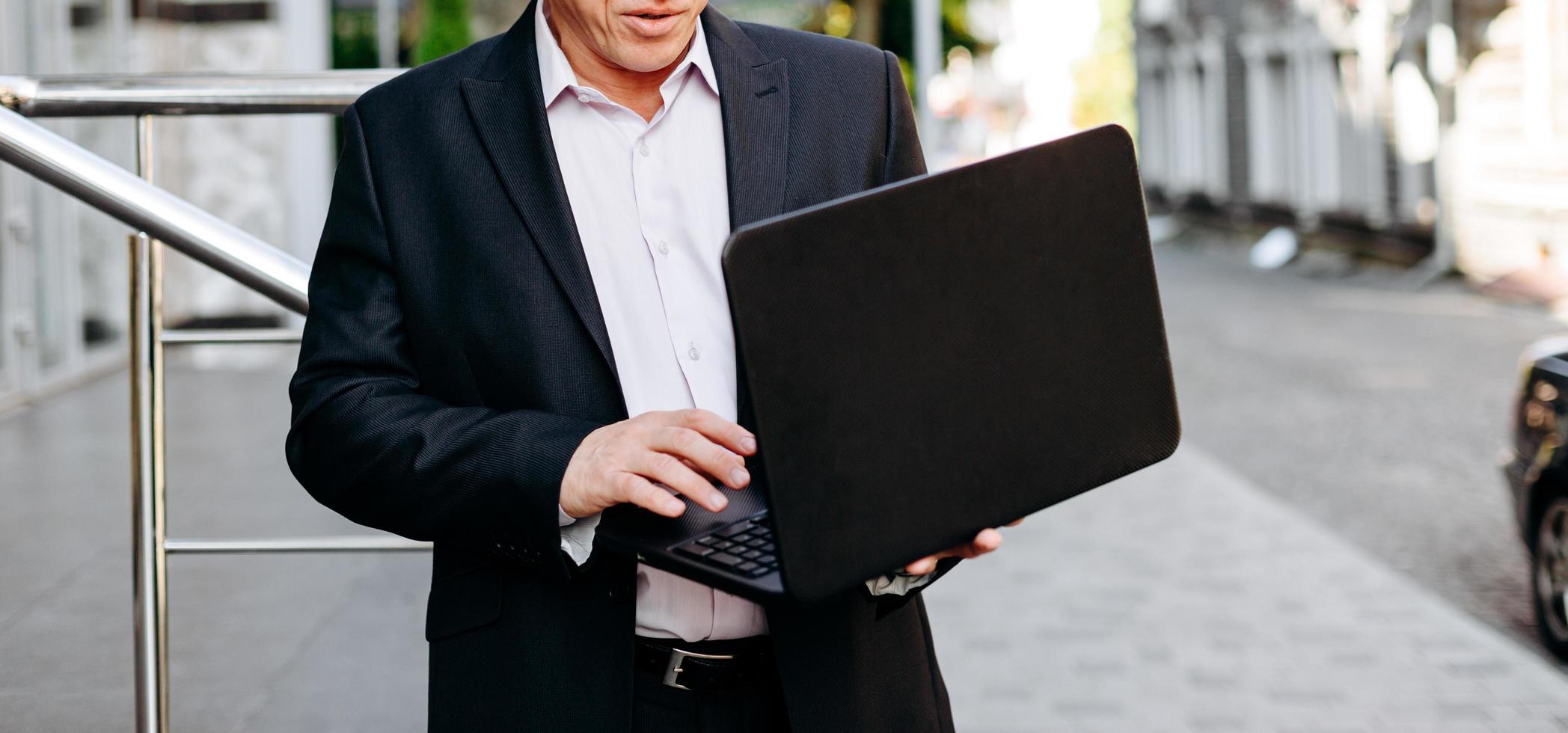 Recortando la imagen del empresario senior sosteniendo una computadora portátil en la mano y escribiendo de pie en la ciudad.- imagen foto