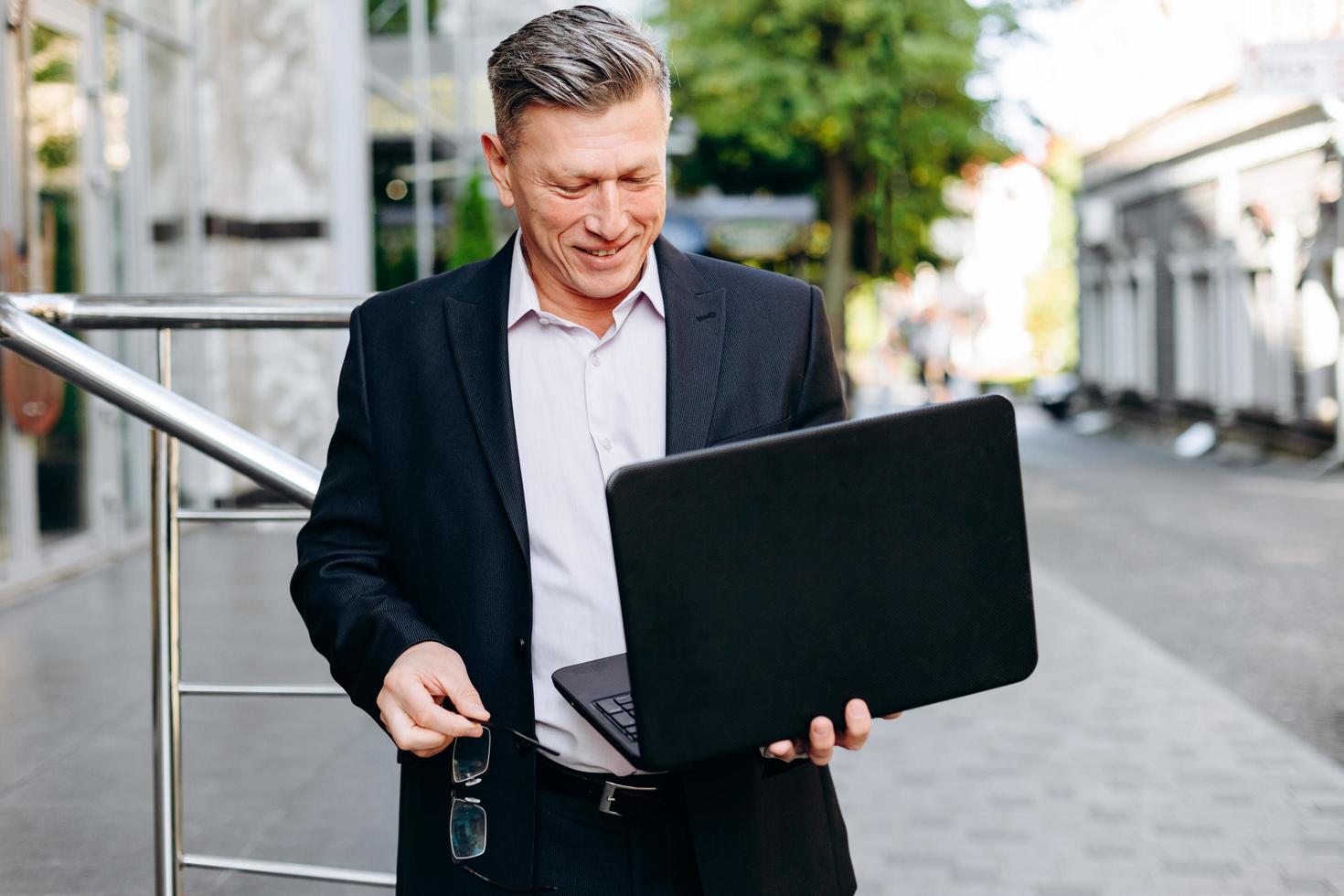Happy senior businessman holding laptop in his hand in city outdoor and looking at the screen.- Image photo