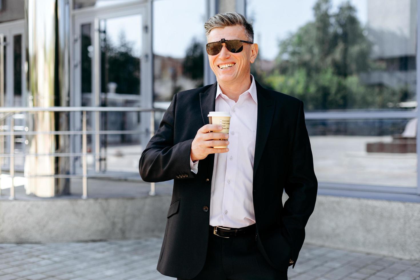 Portrait of  senior businessman standing on pavement holding a cup of coffee and smiling . - Image photo