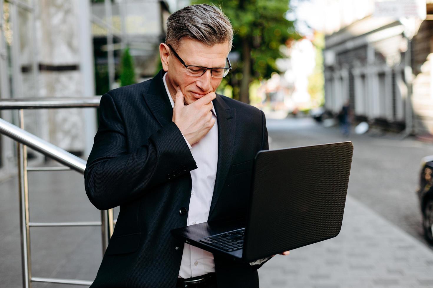 Senior businessman holding open laptop and attentively looking at the screen touches his face- Image photo