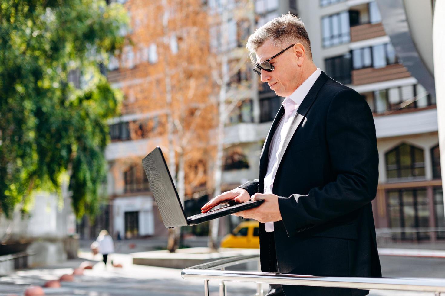 serio empresario senior en gafas de sol sosteniendo una computadora portátil y trabajando en ella al aire libre. - imagen foto