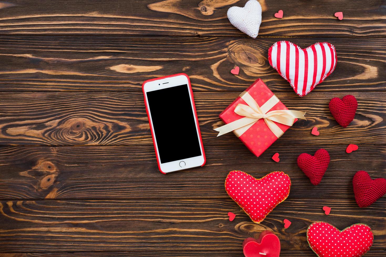 composición linda laicos plana con corazones rojos de tela hechos a mano y teléfono móvil. caja de regalo roja sobre mesa de madera. feliz cumpleaños o felicitaciones de aniversario. plantilla de historia de amor romántico con espacio de copia. foto