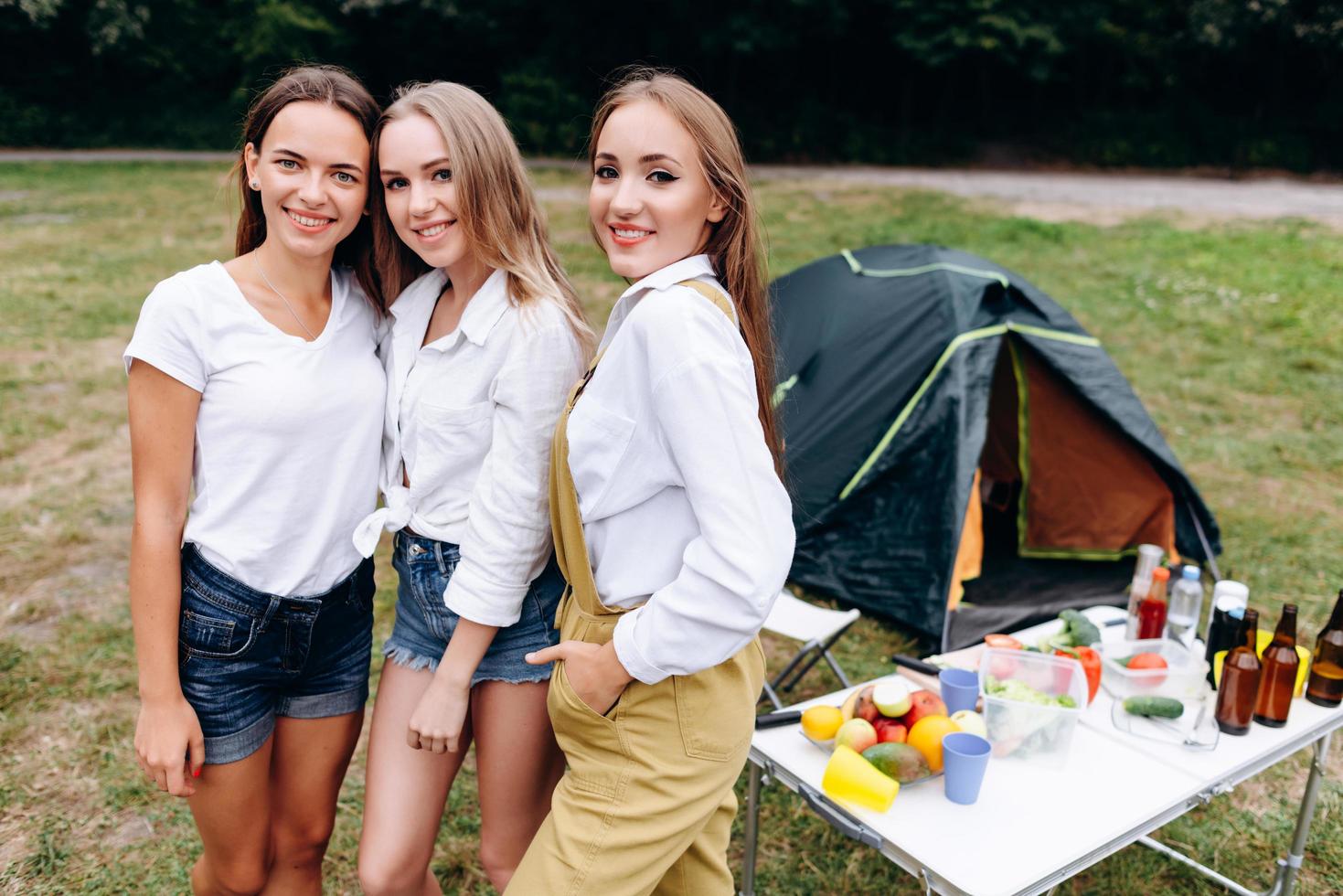 Tres bonitas chicas de pie en el camping y mirando a la cámara. foto