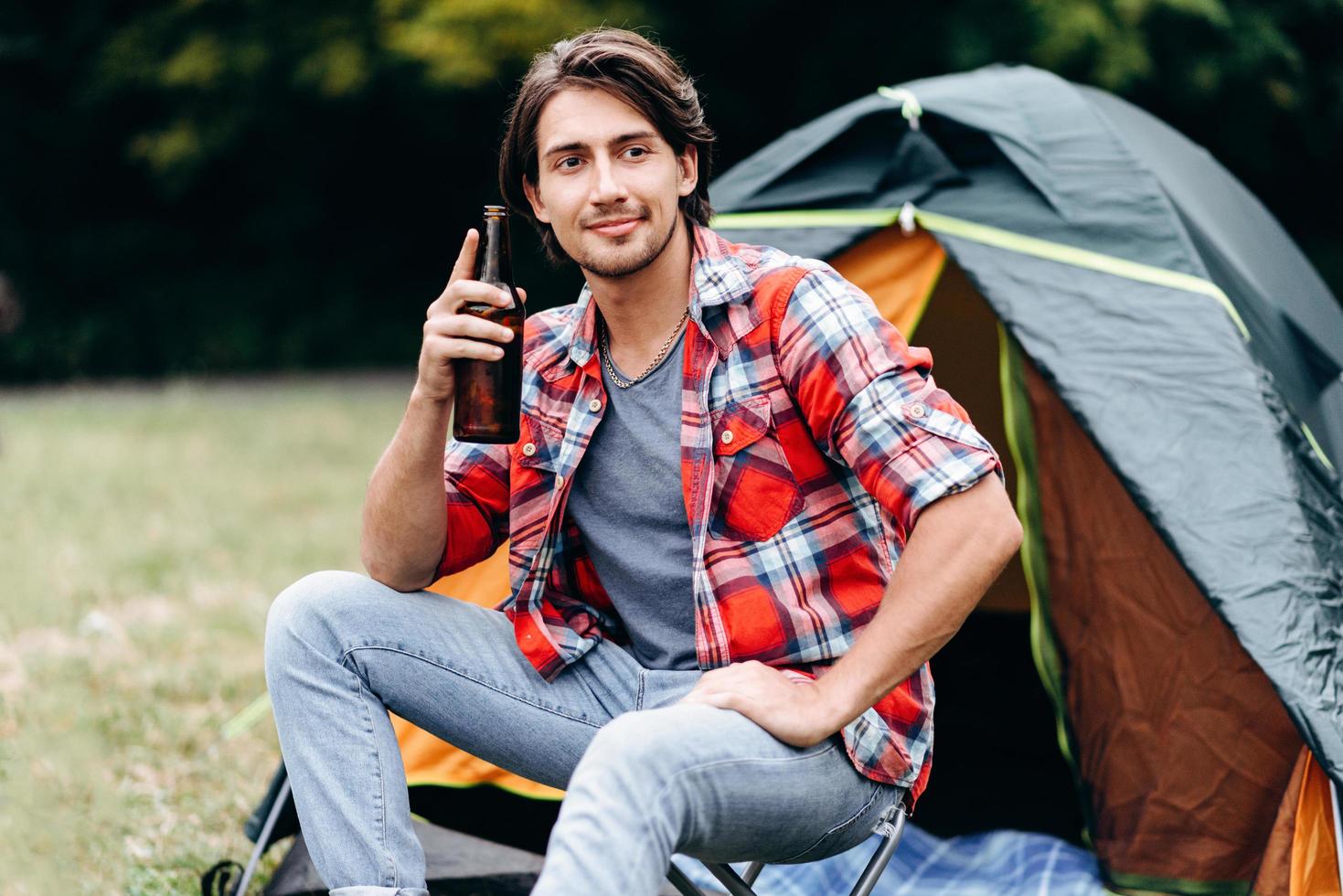 A guy holding a bottle with beer and smiling looking out. Tent on the background photo