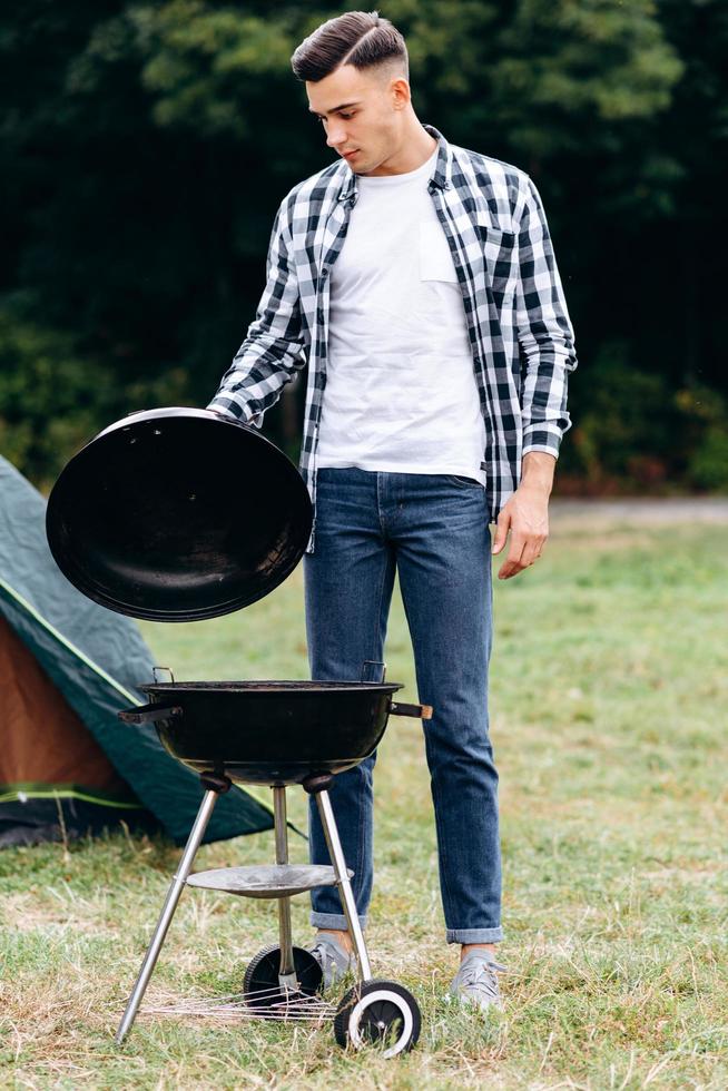 chico joven cocinando en el camping. de pie y abre la tapa de la barbacoa foto