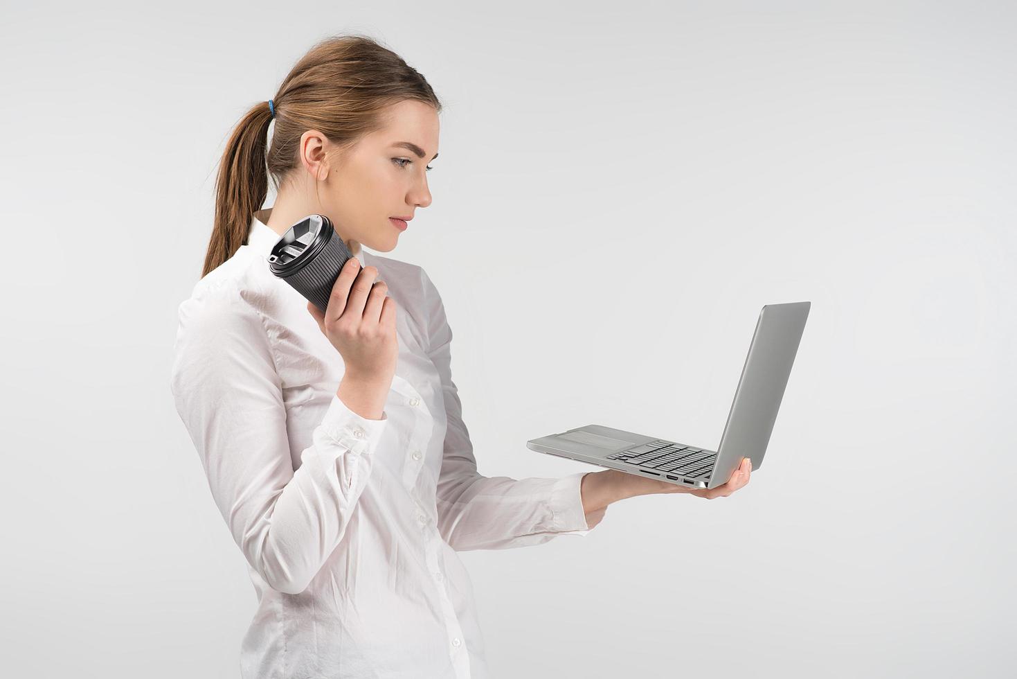 mujer con camisa blanca mirando la computadora portátil. sosteniendo una taza de café y un dispositivo en sus manos mientras está de pie contra el fondo. foto