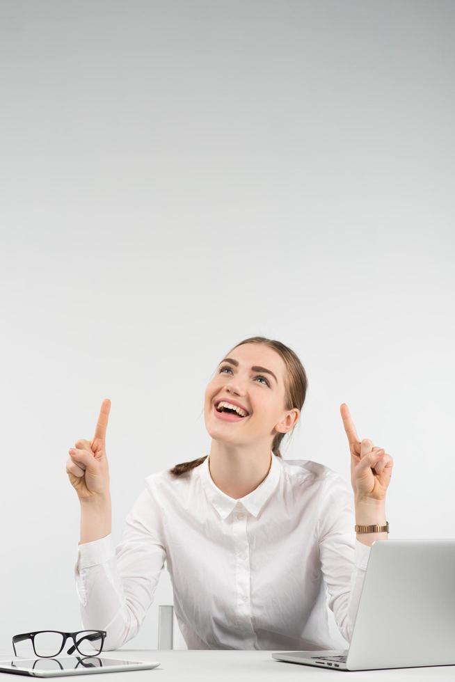 mujer feliz se sienta junto a una computadora portátil y, riendo, mira hacia arriba apuntando con el dedo hacia arriba con ambas manos. - imagen vertical foto