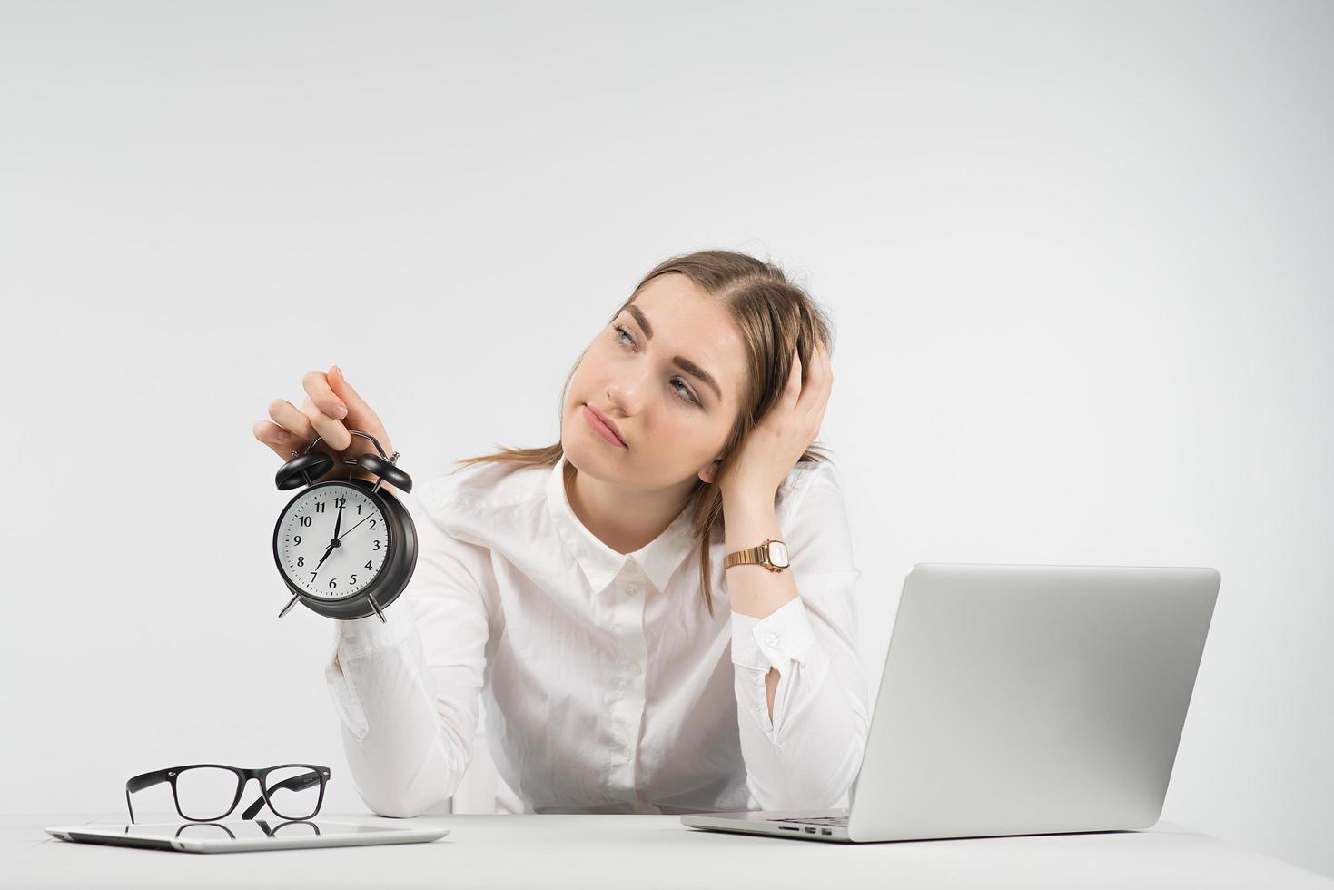 Sad woman sits next a laptop and looks away holds alarm clock and puts her head on the  her arm photo