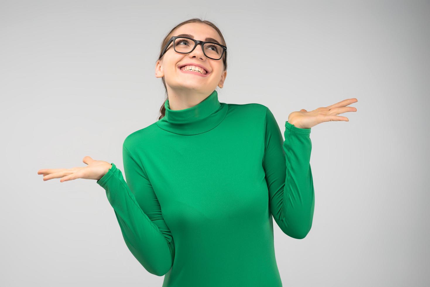 Cheerful young girl in glasses posing in studio shrugs in surprise and looks up photo