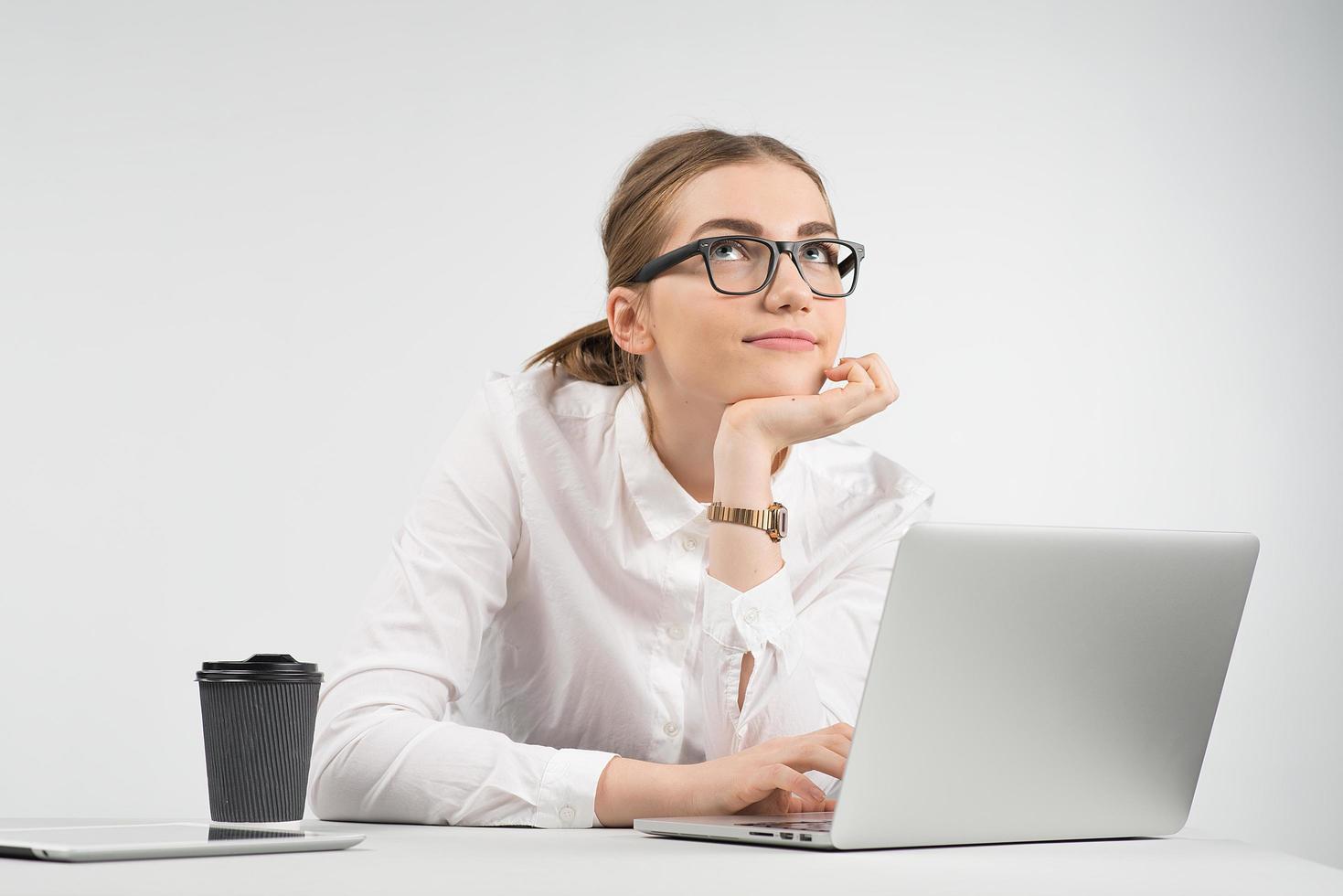 Smiling business woman sitting behind a laptop with a cup of coffee and ipad on the table and looking up dreamy photo