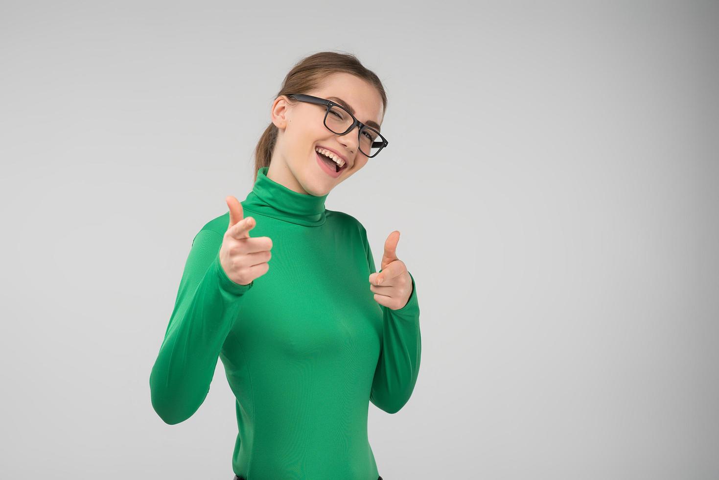 Chica adolescente en ropa casual con gafas guiña felizmente apuntando con el dedo hacia adelante foto