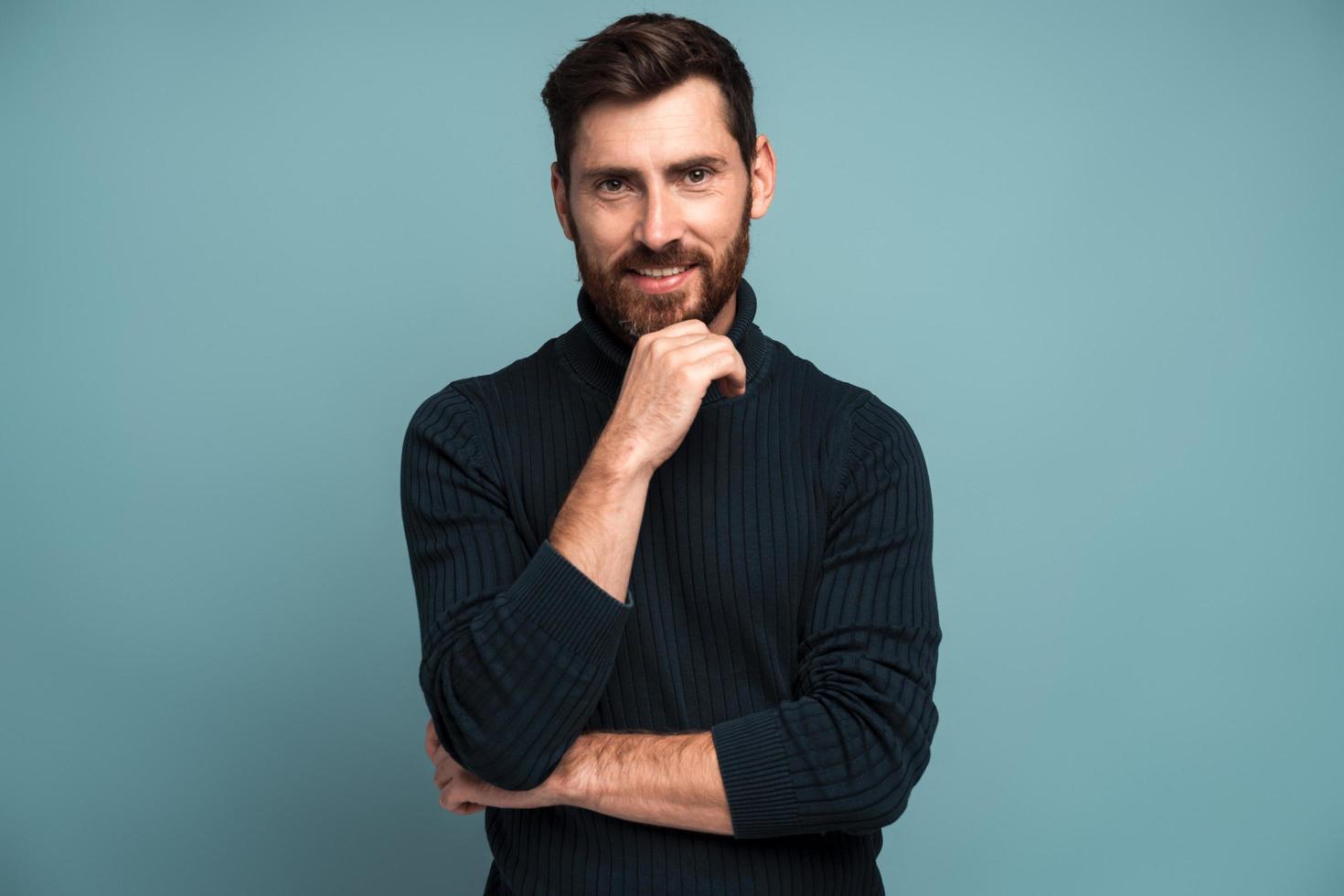 Portrait of happy unshaven man standing with hand near his face and smiling to the camera. Male appearance concept. Indoor studio shot isolated on blue photo