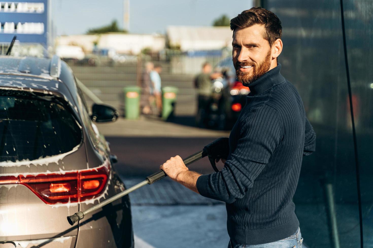 Vista vertical de cintura para arriba del hombre lavando su auto en la calle usando una hidrolimpiadora de alta presión con espuma. placer masculino sonriendo a la cámara foto