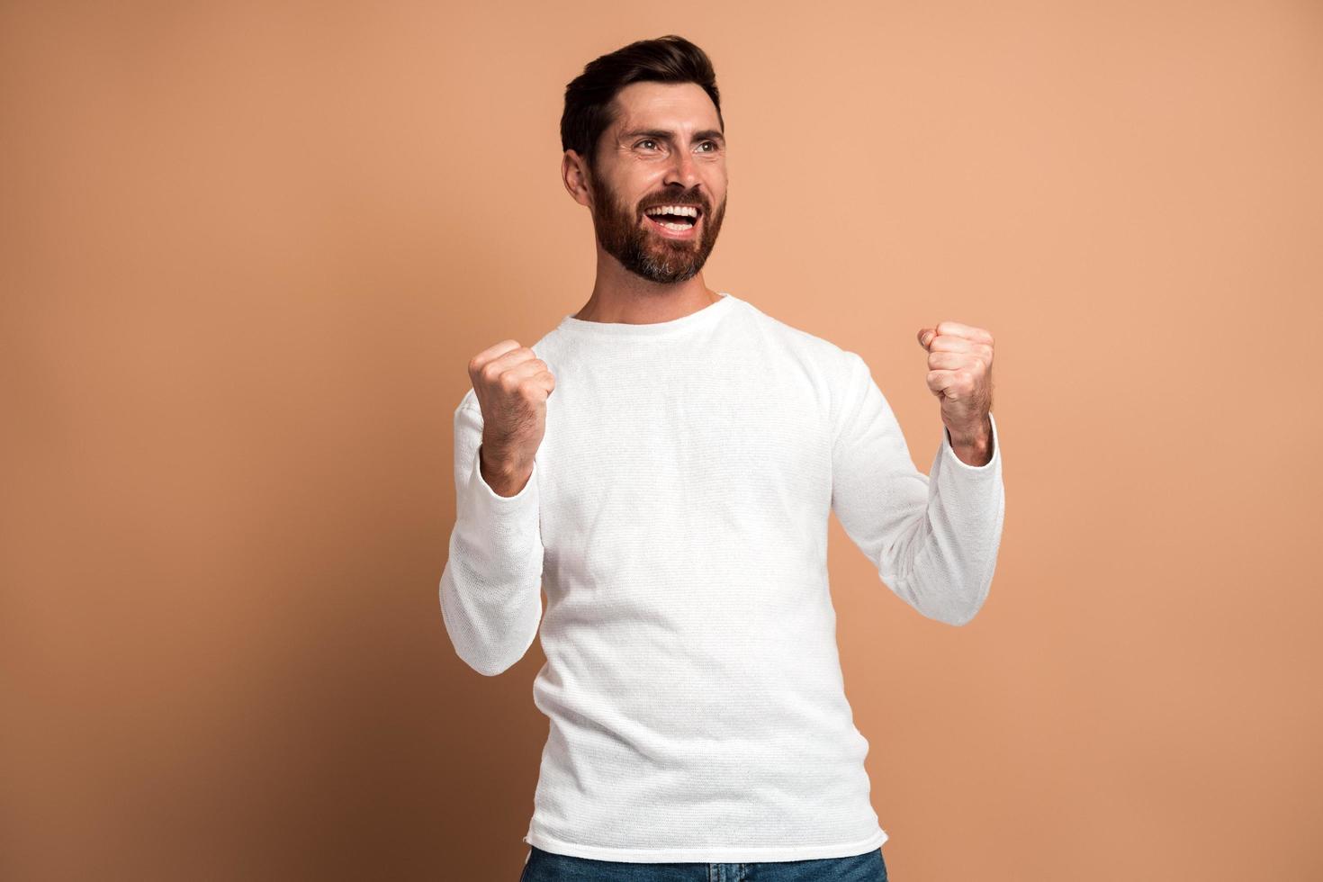 Hombre con barba muy emocionado y lleno de alegría haciendo un gesto de sí, asombrado con su victoria, triunfo. Foto de estudio de interior aislado sobre fondo beige