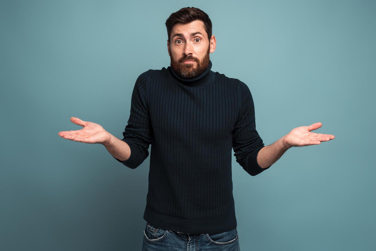 I don't know. Portrait of confused handsome bearded young man standing with raised arms and looking at camera with answer. Indoor studio shot, isolated on blue background photo