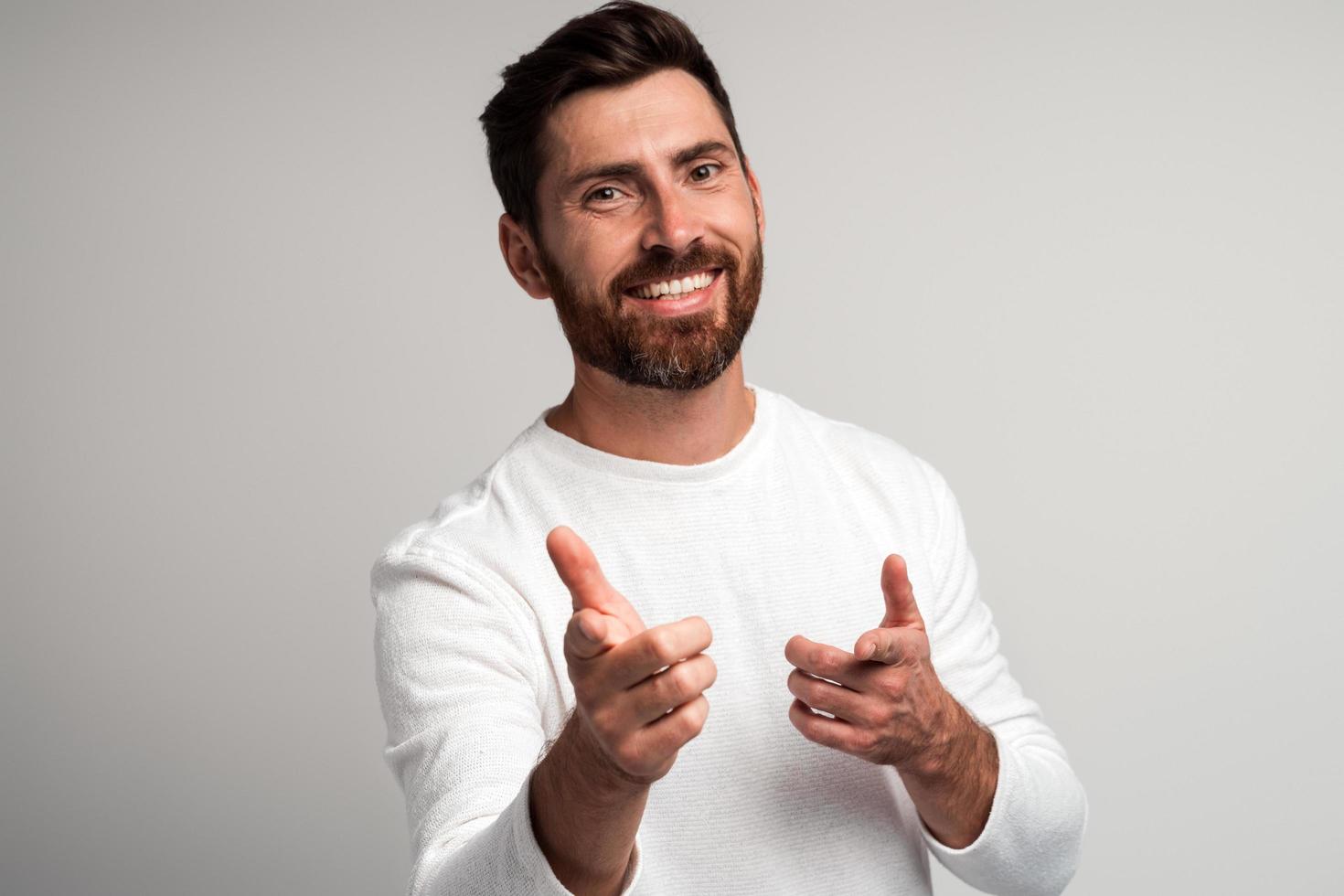 Eh, tú. Retrato de hombre barbudo feliz con camisa blanca sonriendo y apuntando a la cámara sobre fondo gris claro. tiro del estudio foto