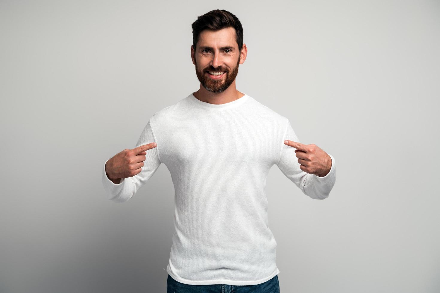 Retrato de hombre engreído con camisa blanca apuntando a sí mismo y mirando con expresión arrogante y egoísta, sintiéndose exitoso y engreído. Foto de estudio de interior aislado sobre fondo blanco.