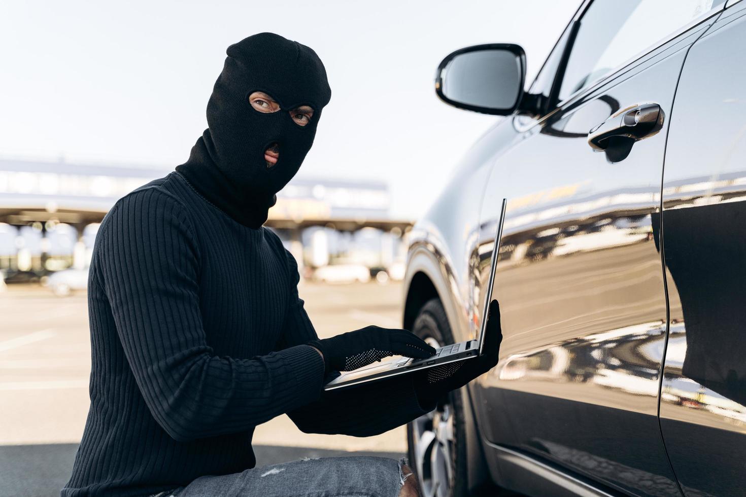 Retrato de cintura para arriba del ladrón de automóviles con sistema de alarma de piratería de computadora portátil mientras está sentado cerca del automóvil durante el día. foto de stock