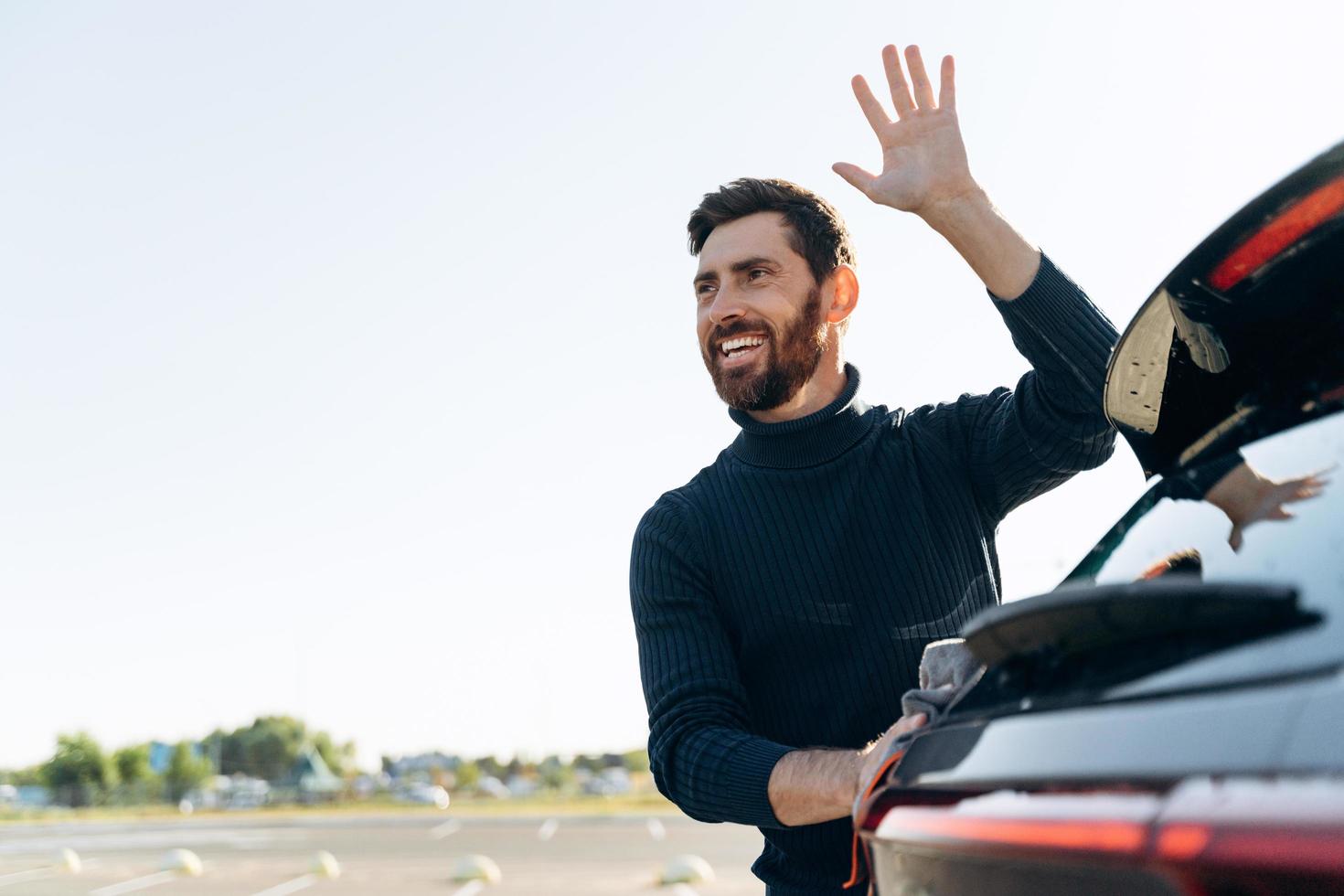 Waist up portrait of the handsome caucasian man holding the microfiber in hand and polishing the car while waving to somebody at the street photo