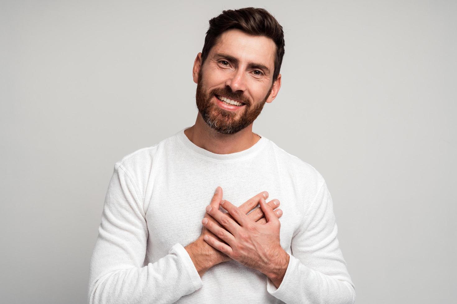 Waist up portrait view of the young caucasian man feeling romantic, happy and in love, smiling cheerfully and holding hands close to heart against white wall photo