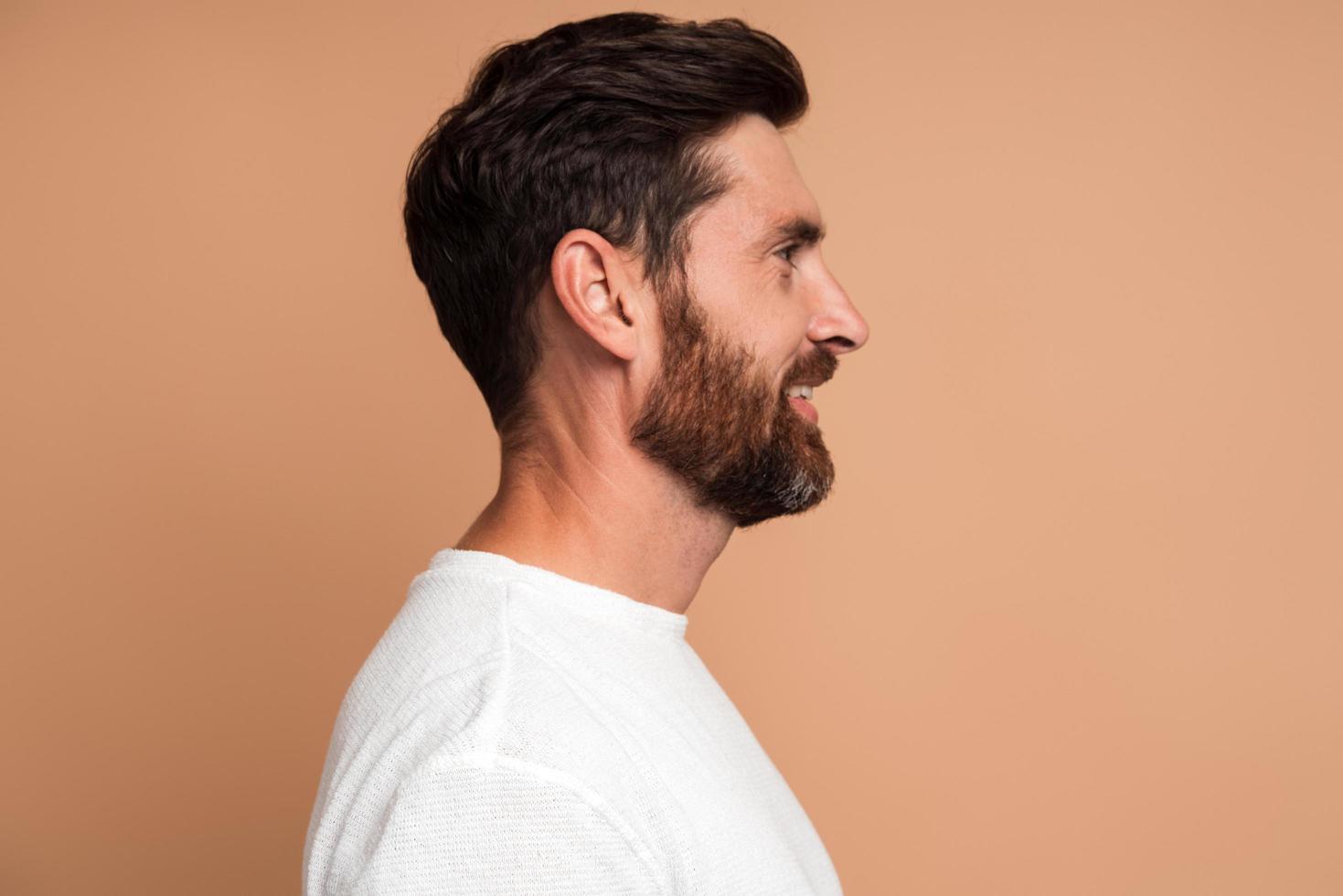 Vista lateral del hombre moreno guapo positivo con barba en camisa blanca de pie con una sonrisa de placer y mirando a otro lado con expresión alegre. Foto de estudio de interior aislado sobre fondo beige