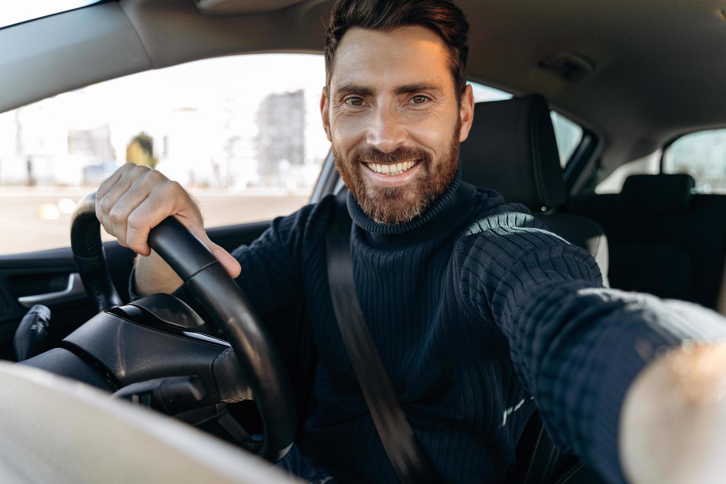 gran selfie. Hombre guapo con barba sosteniendo la cámara y haciendo selfie mientras está sentado en el automóvil en el asiento del conductor y mirando a la cámara con una sonrisa de placer foto