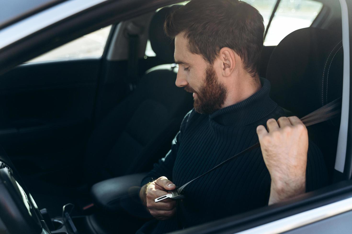 Side view of the caucasian young bearded man in a car fastening his seat belts before the driving. Confident guy riding at the work. Stock photo