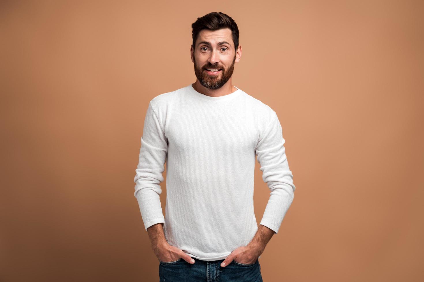 Portrait of brutal handsome bearded young man model in white shirt standing and looking at the camera with pleasure proud face. Indoor studio shot isolated on beige background photo