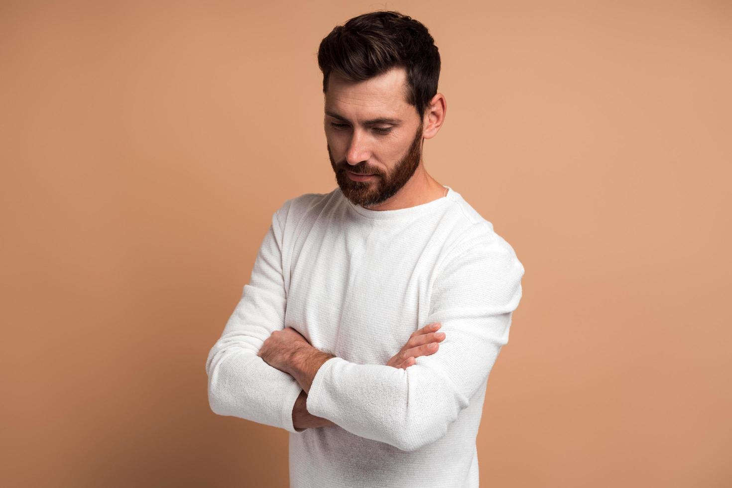 Portrait of handsome pensive bearded young man standing with crossed arms and looking down with pondering expression while posing in front of the camera on beige background photo