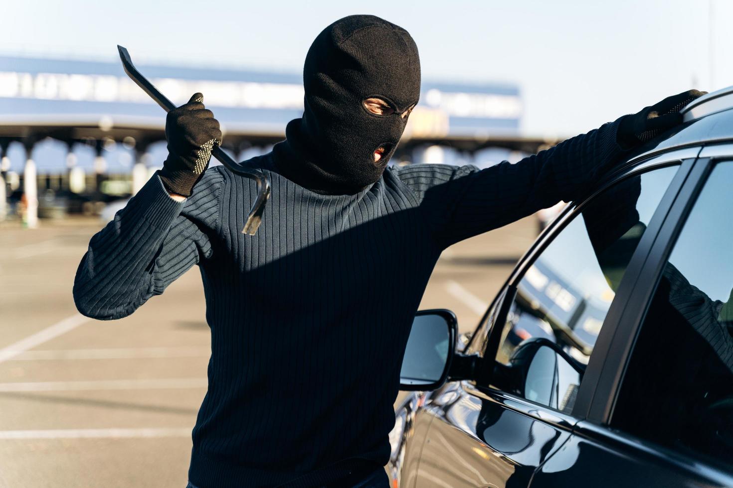 Hombre peligroso vestido de negro con un pasamontañas en la cabeza preparándose para romper con una palanca el cristal del coche antes del robo. ladrón de autos, concepto de robo de autos foto