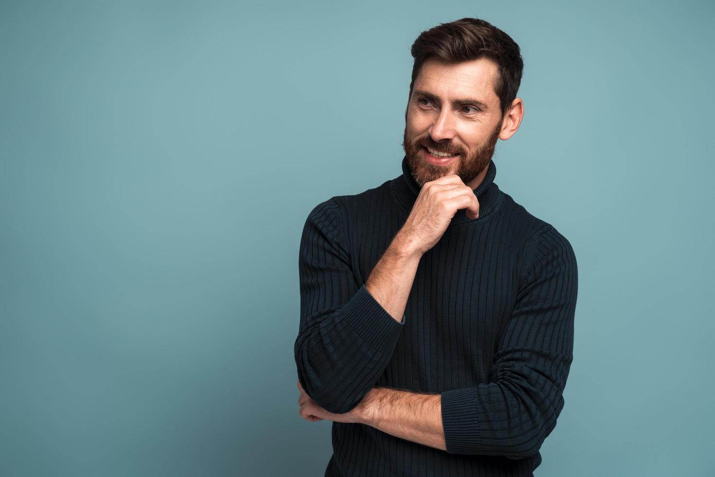 Portrait of cheerful unshaven man standing with hand near his face and smiling while looking away. Male appearance concept. Indoor studio shot isolated on blue photo