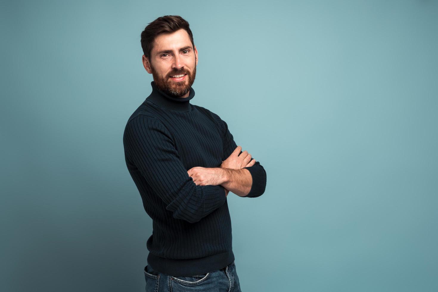 Portrait of cheerful good-natured bearded young man crossing arms and smiling sincerely at camera, looking satisfied contented with life. Studio shot isolated on blue background photo