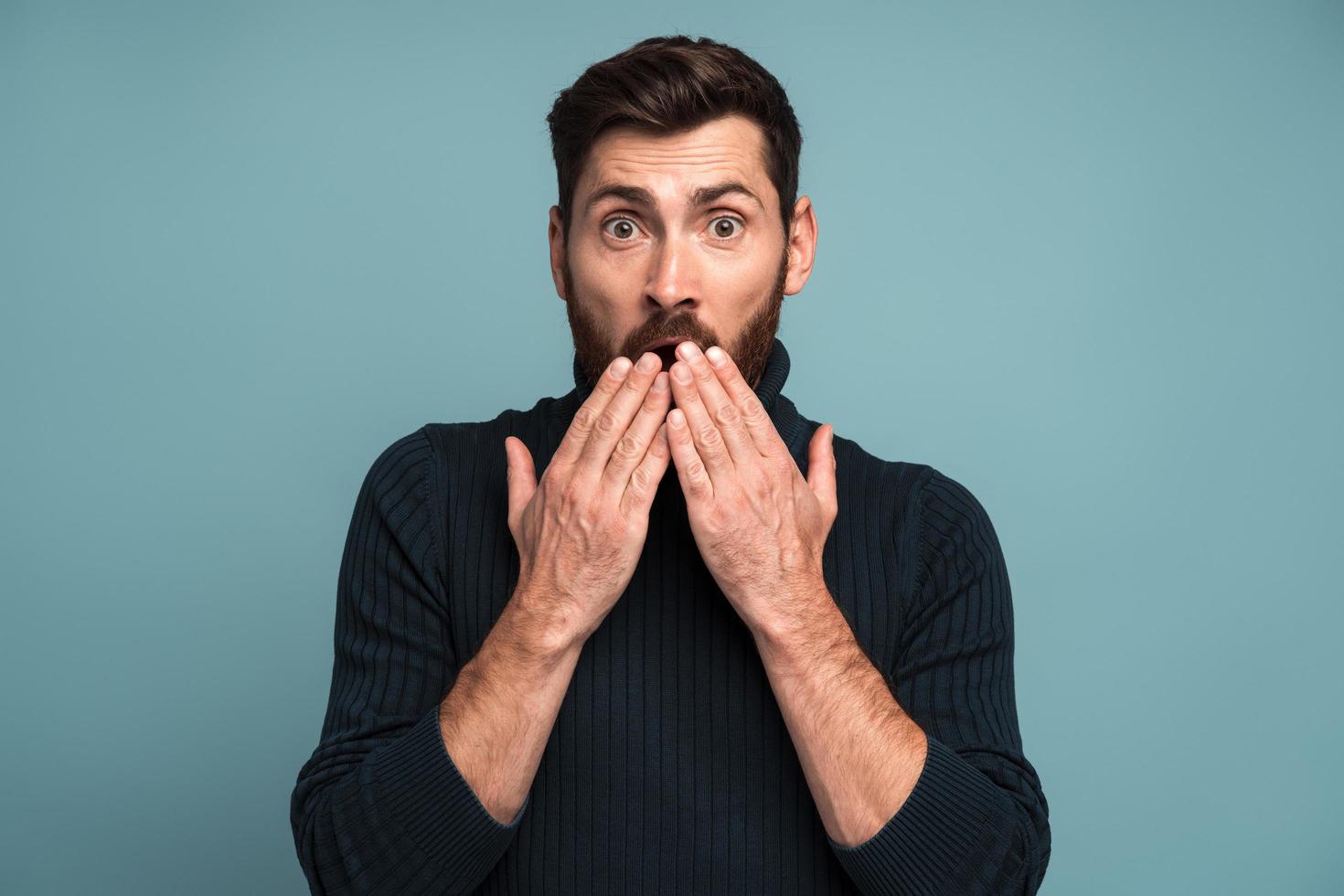 Shocked man closing mouth with hands, confused and depressed with information, looking with astonishment and fear, privacy. Indoor studio shot isolated on blue background photo