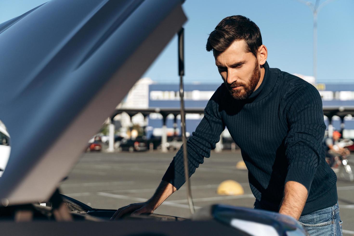 Upset bearded man checking his car engine after breaking down on the road. Serious male is standing in front of the opening hood. Transportation concept photo