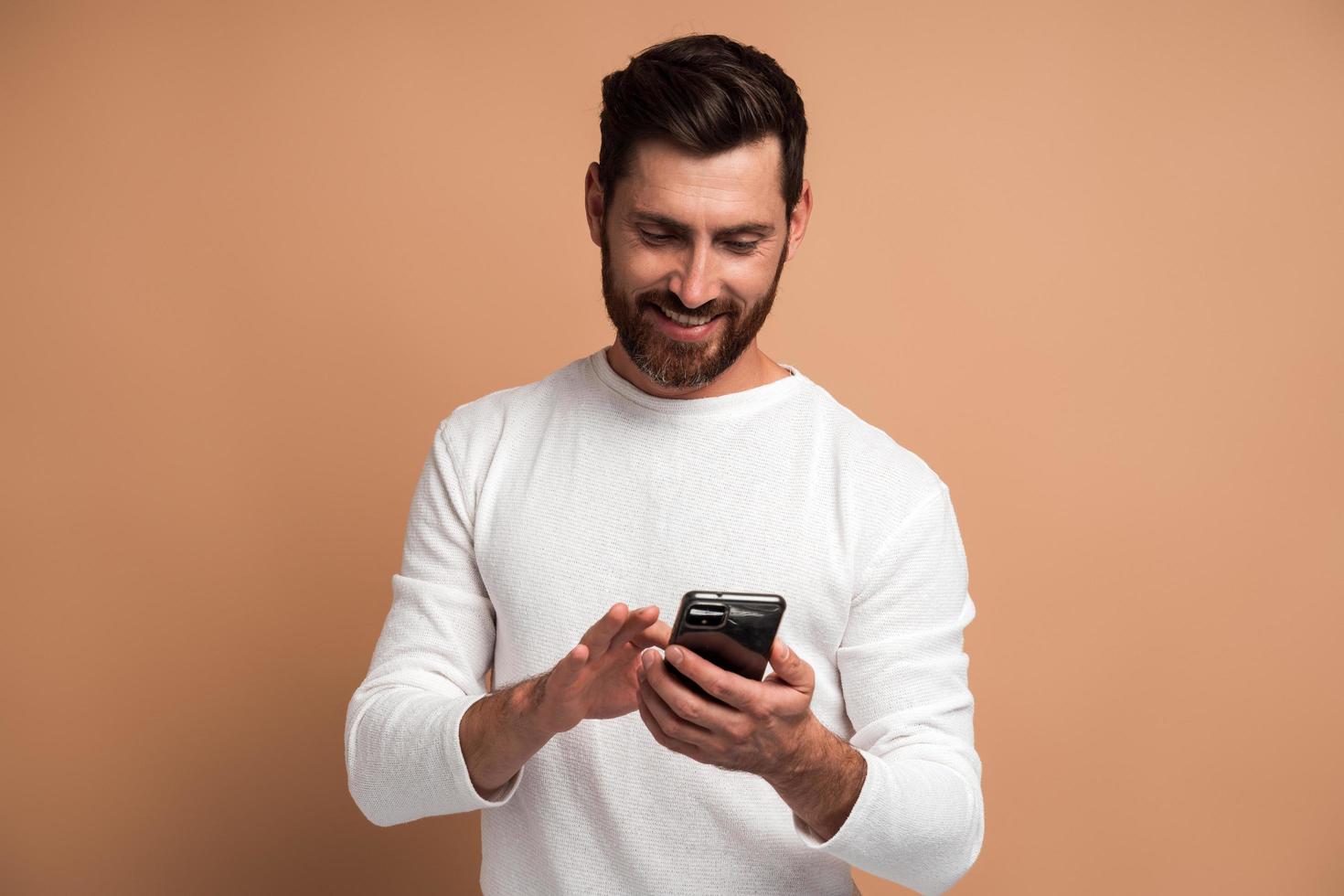 Hombre barbudo asertivo mirando atentamente la pantalla del teléfono inteligente que sostiene, navegando por internet, haciendo compras en línea. Foto de estudio de interior aislado sobre fondo beige